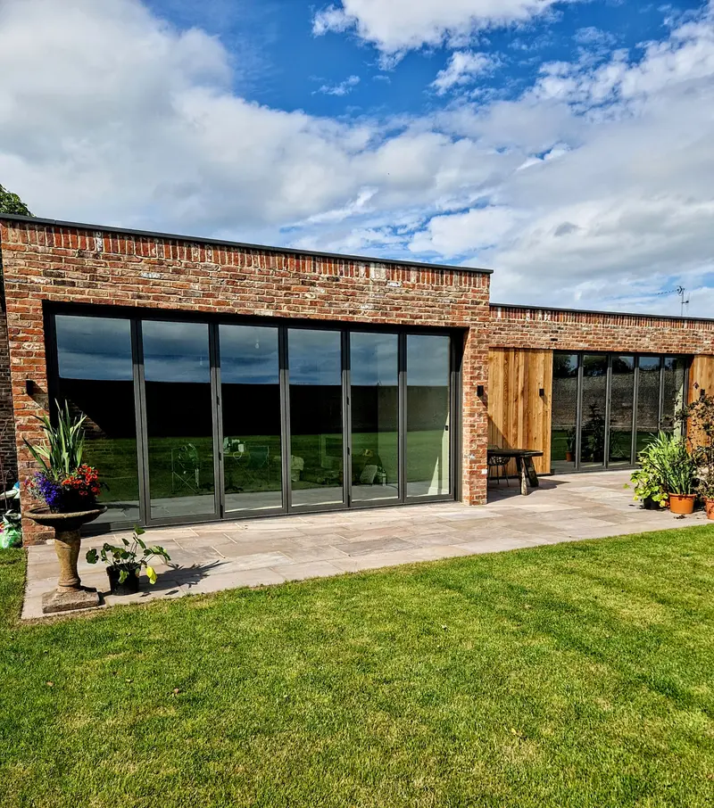 Anthracite aluminium bifolds, closed, in a red brick and timber-clad barn conversion facing a lawn