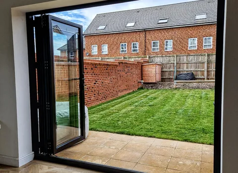 Black bifold folded open from inside onto a stone-paved patio and small lawn, houses beyond