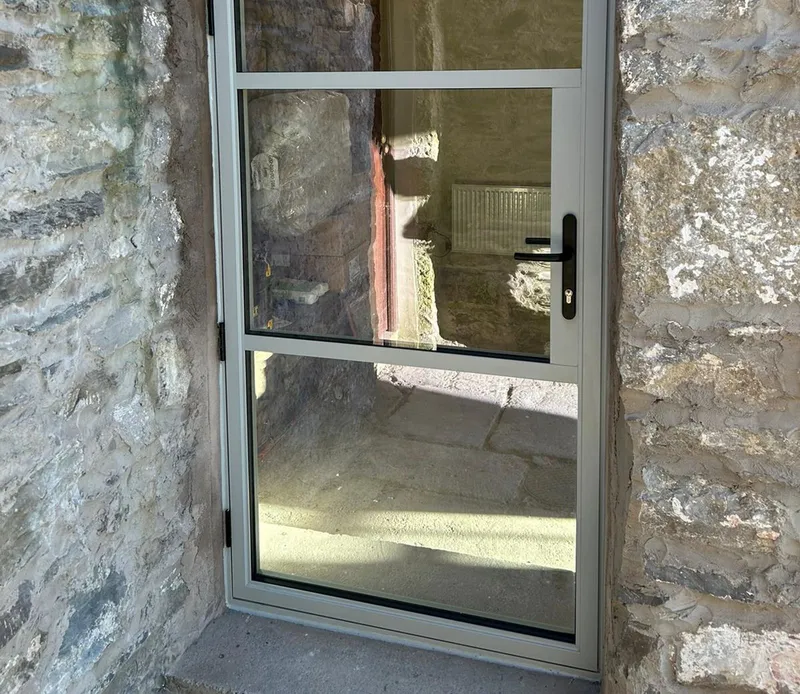 Fully glazed grey aluminium door, closed, set into a rough stone wall; bare stone room beyond