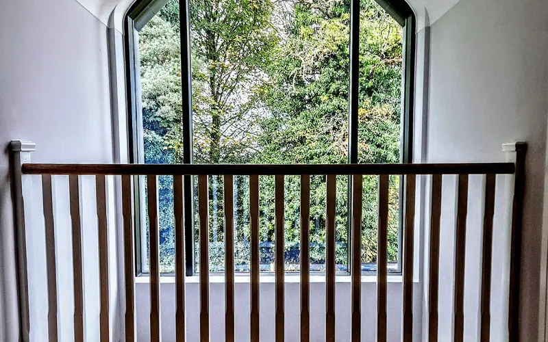 Black gothic-arched window on a stair landing, seen through a dark timber balustrade, trees beyond