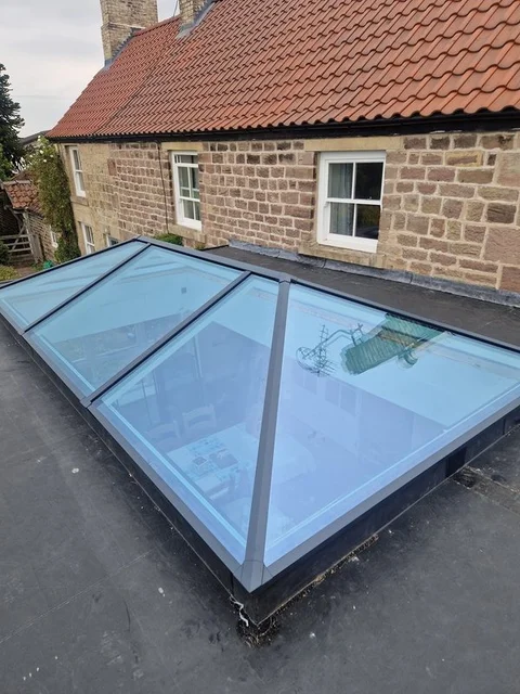 Grey aluminium roof lantern on a flat felt roof, seen from outside, beside a stone cottage with a pantile roof