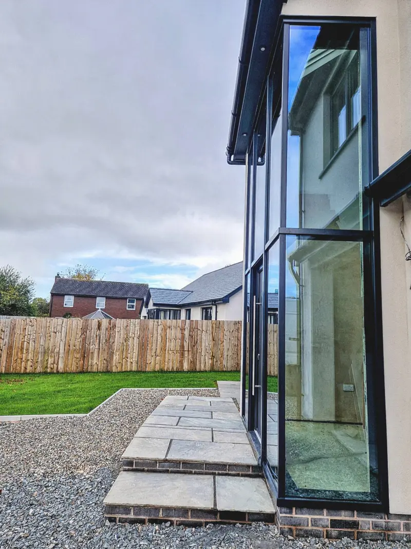 Raking view of a tall anthracite glazed door, closed, on a rendered house above gravel and paving