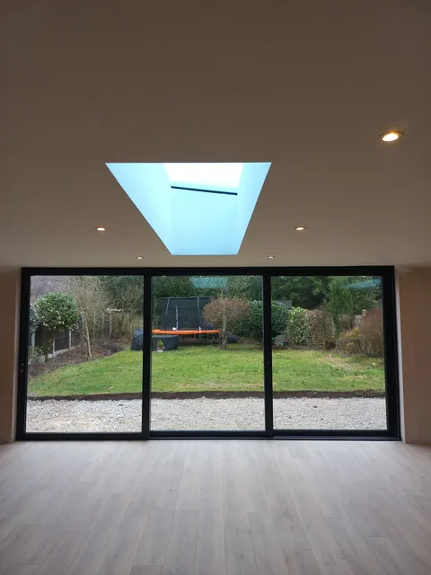 Three-panel black aluminium slider seen from inside an empty room lit by a pyramid rooflight